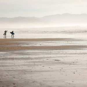 surfers_in_wetsuits_with_surfboards_walk_along_wide_beach_with_tide_out_towards_hazy_water